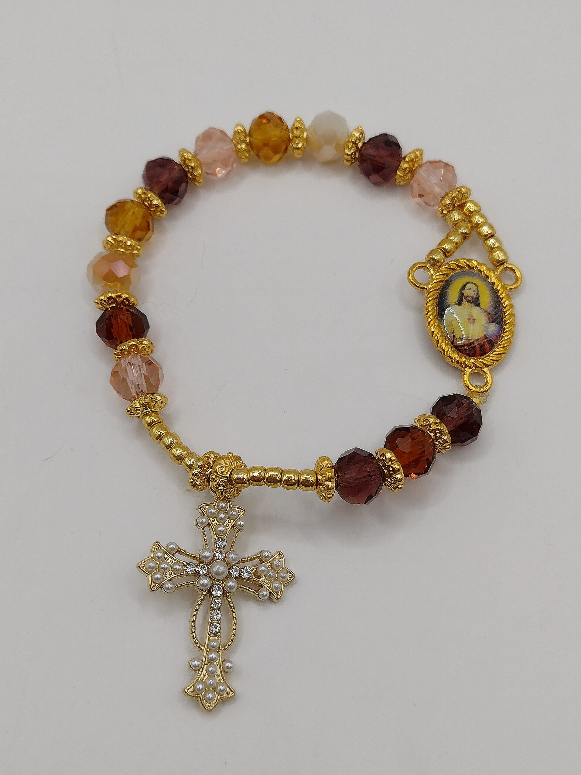 Bransoletka‑różaniec z fasetowanych koralików w kremowo‑brązowych odcieniach, z medalikiem Jezusa i ozdobnym krzyżykiem.
