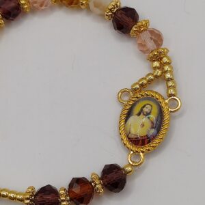Bransoletka‑różaniec z fasetowanych koralików w kremowo‑brązowych odcieniach, z medalikiem Jezusa i ozdobnym krzyżykiem.