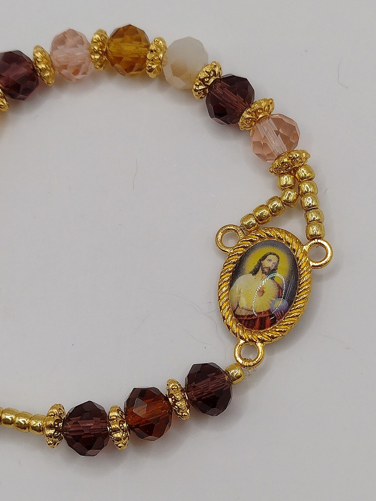 Bransoletka‑różaniec z fasetowanych koralików w kremowo‑brązowych odcieniach, z medalikiem Jezusa i ozdobnym krzyżykiem.