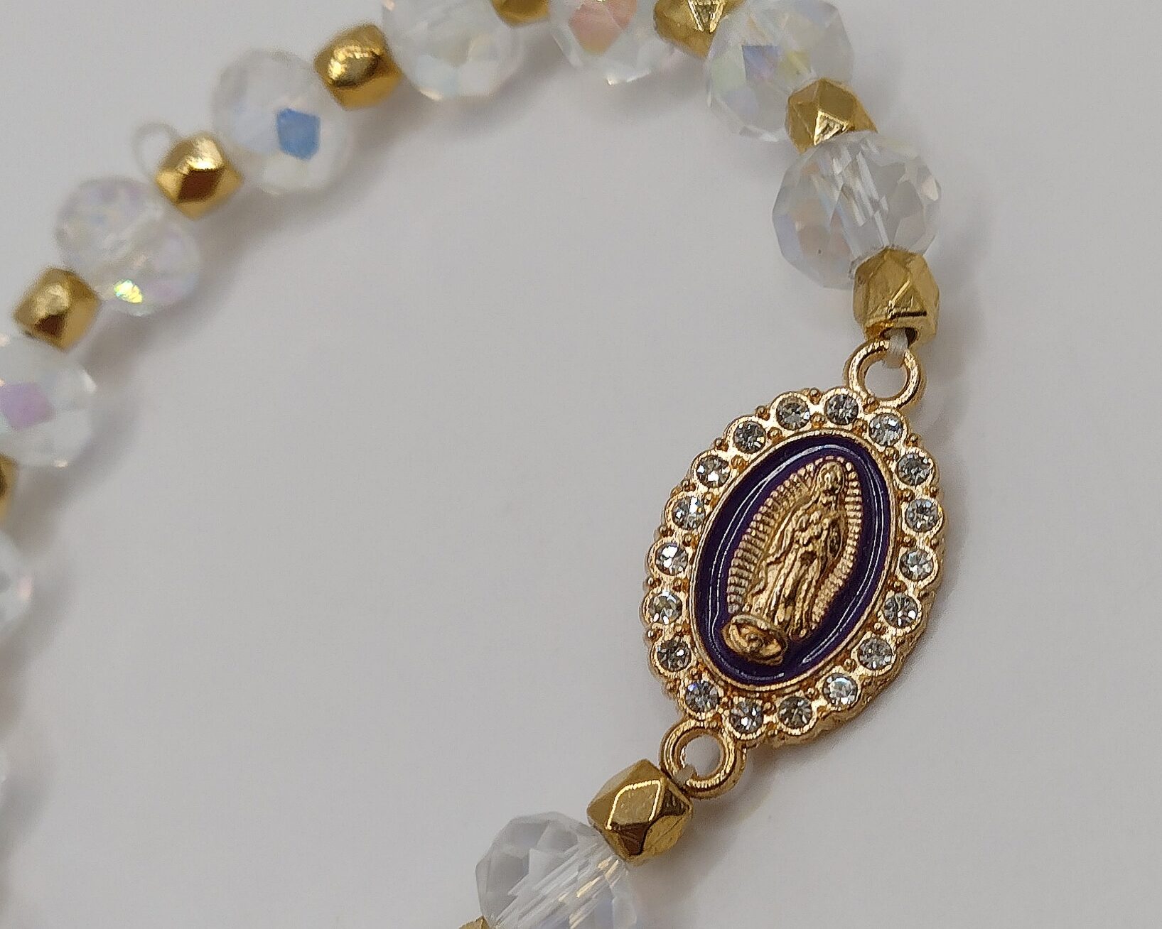 Bransoletka‑różaniec z białych fasetowanych koralików na elastycznej gumce, z ozdobnym krzyżykiem i medalikiem Matki Boskiej.